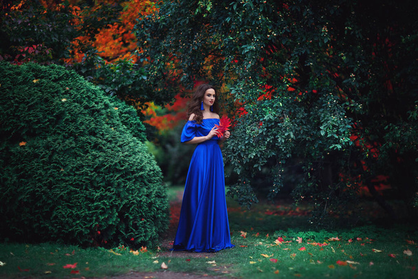 Romantisches Porträt eines schönen jungen Mädchens mit blauem Make-up und roten Lippen, in einem langen ultramarinblauen Kleid mit langen blauen Ohrringen, vor dem Hintergrund einer Herbstarche mit roten Blättern - Foto, Bild
