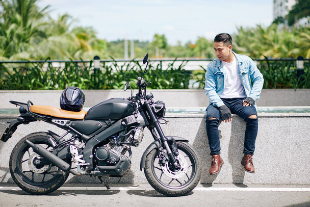 Trots Vietnamese motorrijder zittend op parapet en kijkend naar zijn motorfiets - Foto, afbeelding