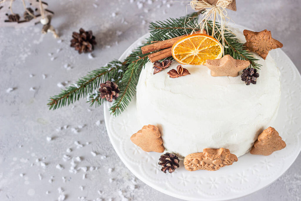 Christmas or New Year gingerbread cake with white cream decorated with gingerbread cookies, fir tree branches, cones, spices and dried orange slice. Winter cake. Grey background. - Photo, image