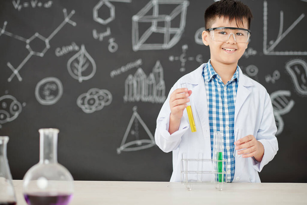Portrait of happy Asian schoolboy mixing colorful liquids in text-tubes in class and looking at camera - Foto, Imagen