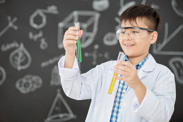 Excited little scientist looking at test-tubes with green and yellow liquids in his hands - 写真・画像