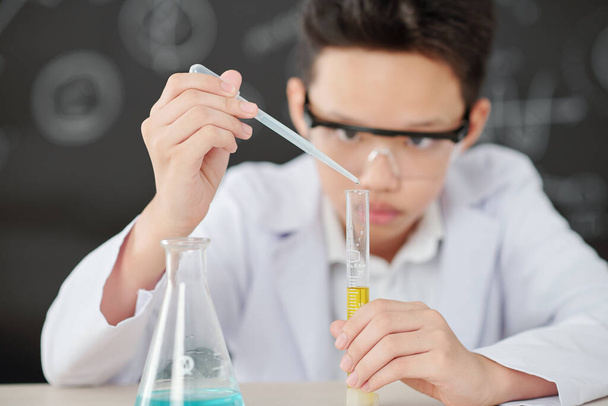 Vietnamese schoolboy adding drop of reagent in test-tube with yellow liquid - Φωτογραφία, εικόνα