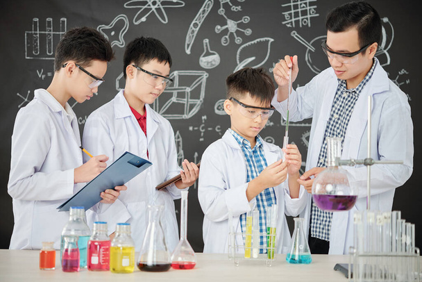 Science leraar helpen student om reagens toe te voegen in test-tube met druppelaar, andere jongens kijken en notities maken - Foto, afbeelding