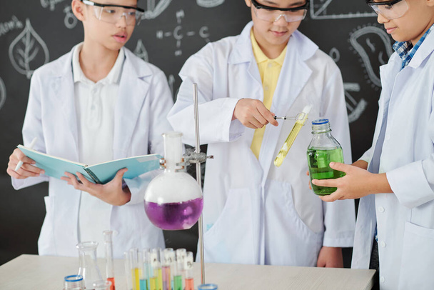 Καλλιεργημένη εικόνα μαθητών σε labcoats που προσθέτουν αντιδραστήριο σε μεγάλο ποτήρι όταν ο συμμαθητής τους γράφει αποτελέσματα πειραμάτων σε βιβλίο - Φωτογραφία, εικόνα