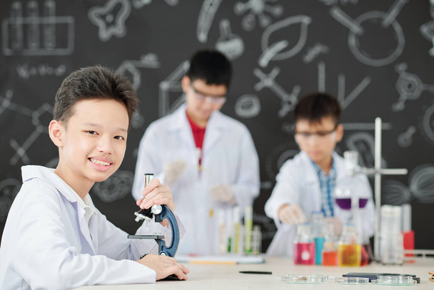 Pozitivní vietnamské školy student sedí u stolu s mikroskopem, když jeho spolužáci míchání chemických činidel v pozadí - Fotografie, Obrázek