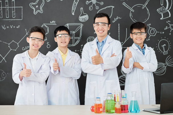Cheerful science teacher and group of school students in labcoats showing thumbs-up and looking at camera - Photo, Image