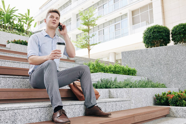 Genç ve mutlu girişimci basamaklarda oturuyor, kahve içiyor ve telefonla arıyor. - Fotoğraf, Görsel