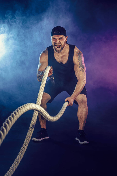 Bearded athletic looking bodybulder work out with battle rope on dark studio background with smoke. Strength and motivation - Photo, Image