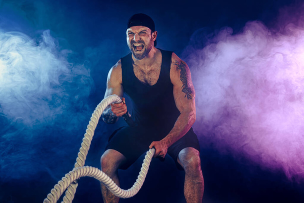 Bearded athletic looking bodybulder work out with battle rope on dark studio background with smoke. Strength and motivation - Foto, Imagen
