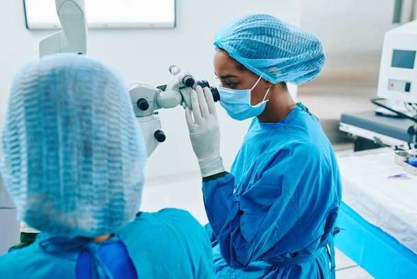 Молодая чёрная женщина-офтальмолог в медицинской маске смотрит через хирургический микроскоп - Фото, изображение