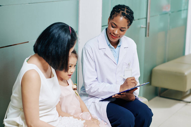 Усміхнена молода педіатр розмовляє з маленькою дівчинкою і її матір'ю і робить нотатки в медичній картці
 - Фото, зображення