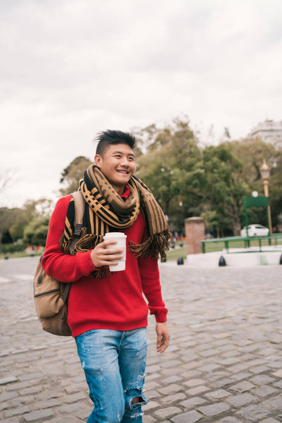 Portrait of young Asian man drinking a cup of coffee while walking outdoors in the street. Urban concept. - Фото, зображення