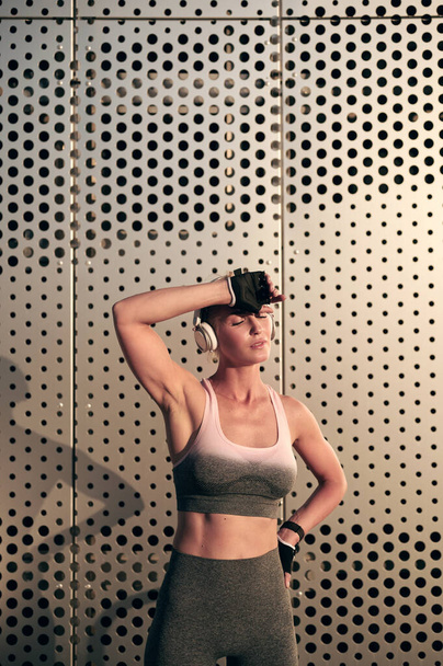 Sterke gespierde fitnessvrouw in sportkleding rustend na de training voor de grijze muur en luisterend naar muziek in een hoofdtelefoon - Foto, afbeelding