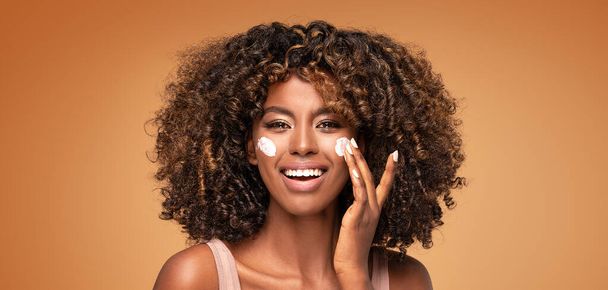 Young afro woman with moisturizer on face. Beautiful african girl applying a cream on cheek. Beauty girl with perfect and healthy skin. - Foto, afbeelding