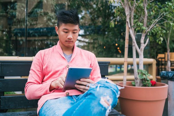 Portrait of young Asian man using his digital tablet while sitting in a bench outdoors. Technology concept. - Fotografie, Obrázek