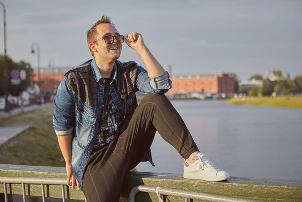 Güneş gözlüklü gülümseyen adam, Rusya 'nın tarihi merkezi Saint-Petersburg' daki köprünün korkuluğunda oturuyor.. - Fotoğraf, Görsel