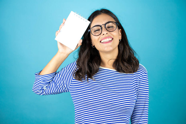 Pretty woman wearing glasses standing over insolated blue background smiling and showing blank notebook in her hand. - Foto, immagini