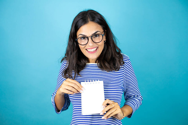 Mulher bonita usando óculos de pé sobre fundo azul insolado sorrindo e mostrando caderno em branco em sua mão. - Foto, Imagem