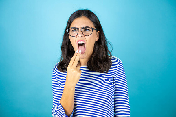 Jolie femme portant des lunettes debout sur fond bleu insolé dégoûté avec sa main à l'intérieur de sa bouche. - Photo, image