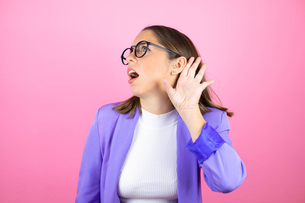 Jovem bela mulher de negócios sobre isolado fundo rosa surpreso com mão sobre orelha ouvindo uma audição para rumor ou fofocas - Foto, Imagem