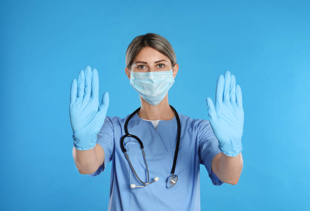 Doctor in protective mask showing stop gesture on light blue background. Prevent spreading of coronavirus - Foto, Imagen