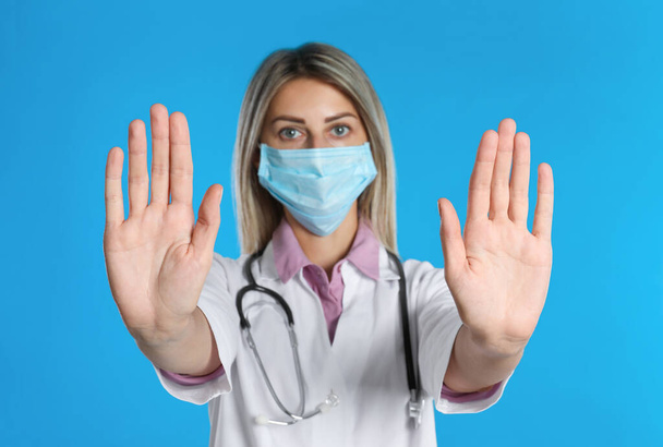 Doctor in protective mask showing stop gesture on light blue background. Prevent spreading of coronavirus - Fotoğraf, Görsel