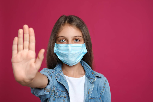 Little girl in protective mask showing stop gesture on crimson background, space for text. Prevent spreading of coronavirus - Foto, Bild