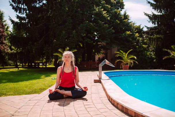 Een blanke vrouw van middelbare leeftijd met vlechten beoefent yoga in de natuur bij het zwembad in een zittende positie met gekruiste benen - Foto, afbeelding