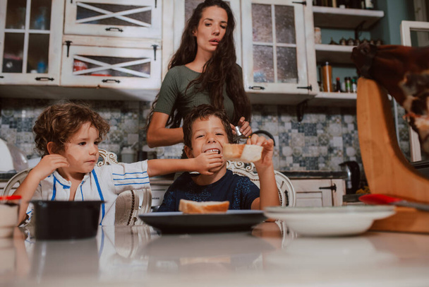 Красива біла мати і двоє синів снідають за столом на кухні. Мати і діти на кухні
 - Фото, зображення