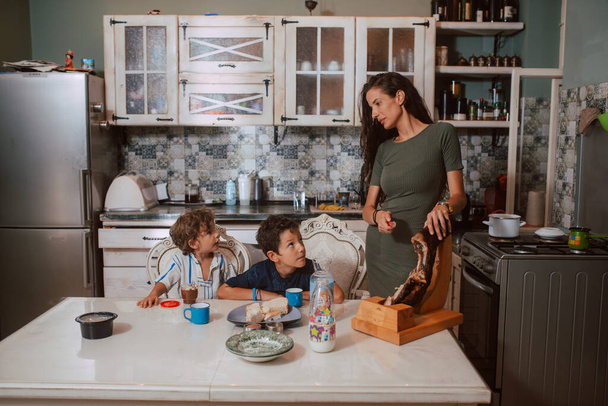 Μια όμορφη λευκή μητέρα με πράσινο φόρεμα ετοιμάζει πρωινό για τους δύο γιους της που κάθονται στο τραπέζι και τρώνε πρωινό στην κουζίνα. Οικογενειακός χρόνος - Φωτογραφία, εικόνα