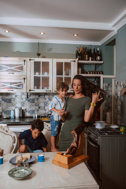 Een mooie blanke moeder in een groene jurk bereidt het ontbijt voor haar twee zoons die aan tafel zitten en ontbijten in de keuken. Gezinstijd - Foto, afbeelding