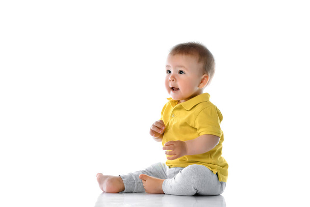 pequeño bebé en un polo amarillo se sienta en el suelo, se ríe, toca sus piernas y mira a un lado en la esquina superior sobre un fondo blanco. - Foto, imagen