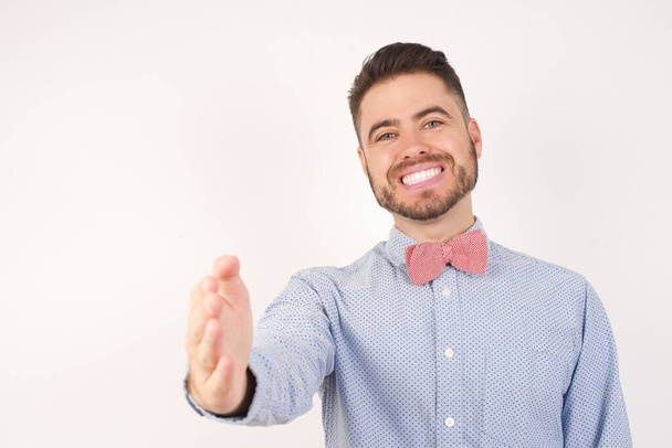 European man dressed in formal shirt and bow tie poses against white studio background smiling friendly offering handshake as greeting and welcoming. Successful business. - Photo, Image