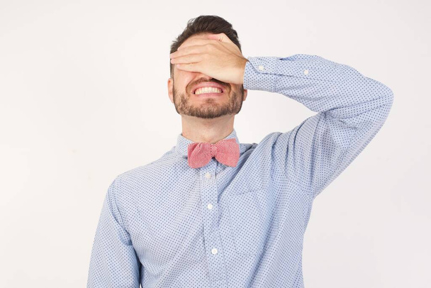 Europäischer Mann in formalem Hemd und Fliege posiert vor weißem Studiohintergrund lächelnd und lachend mit der Hand auf dem Gesicht, die Augen zur Überraschung verdeckt. Blindes Konzept. - Foto, Bild