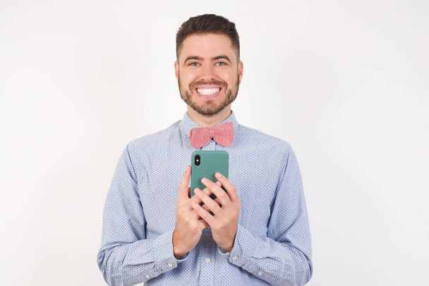 Caucasian man in white studio, wearing bow tie, holding mobile phone, surfs fast unlimited internet, has pleasant smile,  - Photo, Image