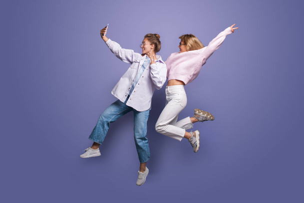 Zwei charmante Frauen in heller Kleidung und blonden Haaren springen auf eine violette Studiowand und machen ein Selfie mit dem Handy - Foto, Bild