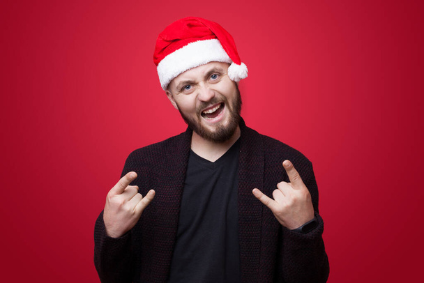 Excited caucasian man with santa hat gesturing rock and roll sign on a red studio wall - Фото, зображення