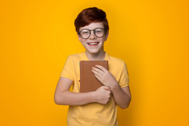 Nahaufnahme eines Ingwerjungen, der ein Buch umarmt und auf einer gelben Studiowand eine Brille trägt, die in die Kamera lächelt - Foto, Bild
