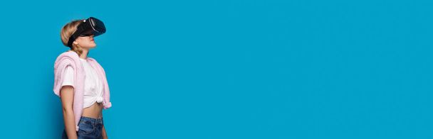 Πλάγια άποψη φωτογραφία μιας ξανθιάς γυναίκας με vr ακουστικά κοιτάζοντας το μπλε banner στούντιο με ελεύθερο χώρο - Φωτογραφία, εικόνα