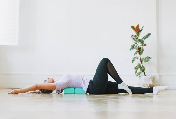 Jovem mulher de uniforme esportivo fazendo alongamento em blocos de fitness deitado no chão em estúdio brilhante - Foto, Imagem