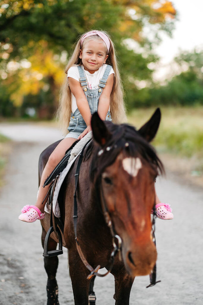 Маленька блондинка з довгим волоссям їде на коні в парку на заході сонця восени. Осінній кінь їде. Дружба дівчини і коня
. - Фото, зображення