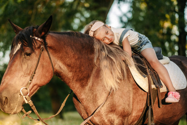 Mała blondynka z długimi włosami kłamie i przytula się na koniu w parku o zachodzie słońca jesienią. Jesienna przejażdżka konna. Przyjaźń dziewczyny i konia. - Zdjęcie, obraz