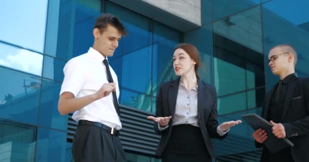 Close up three successful business people stand outdoors near blue glass wall, negotiate, debate, argue. Young confident woman explains to two men knowledge information, actively moves waving hands - Footage, Video