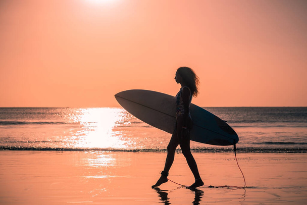 バリのカラフルな夕日でサーフボードとビーチで美しい体を持つ女性サーファーの肖像画 - 写真・画像