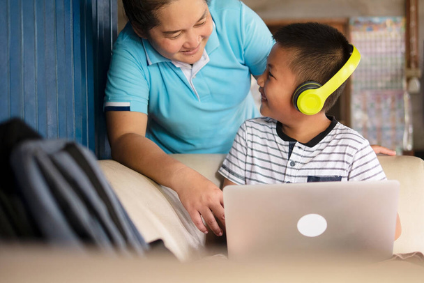 Schulkind Asiatischer Frühchen-Junge mit Kopfhörer und Laptop-Computer, der zusammen mit seiner Mutter in der ländlichen Heimat lächelt, Online-Fernunterricht und Hausaufgabenbetreuung während der COVID-19-Pandemie.  - Foto, Bild