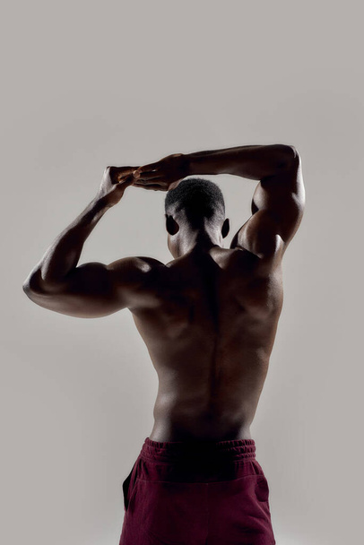 Rear view of a young muscular african american bodybuilder raising his arms while posing shirtless isolated over grey background. Sports, workout, bodybuilding concept - Foto, Imagen