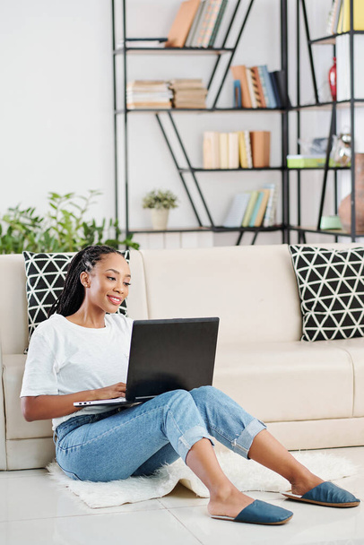 Pozitivní mladá černoška s dredy sedí na podlaze v obývacím pokoji a kódování na notebooku - Fotografie, Obrázek