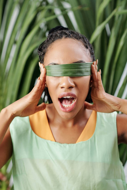 Black young woman covering eyes with palm leaf and opening mouth - Photo, Image