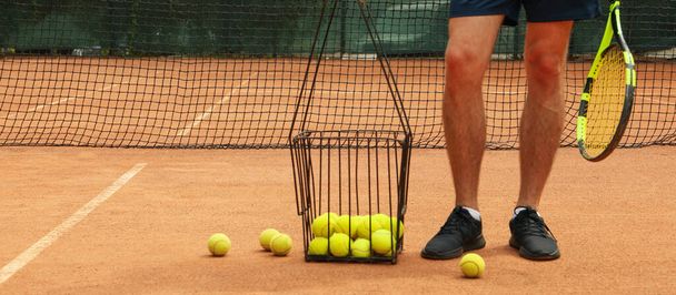 Man hold racket on clay court with basket of tennis balls - 写真・画像
