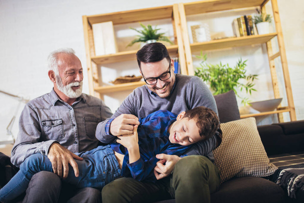 Happy grandfather and father tickling little boy, three generations of men having fun together, sitting on couch. - Photo, Image